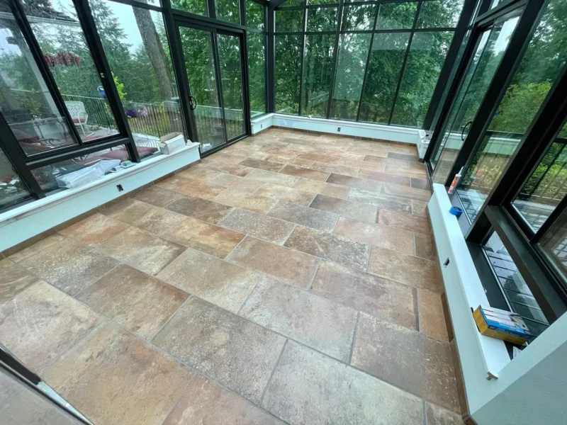 Sunroom or enclosed patio with large black-framed windows and warm stone-look tile flooring overlooking trees