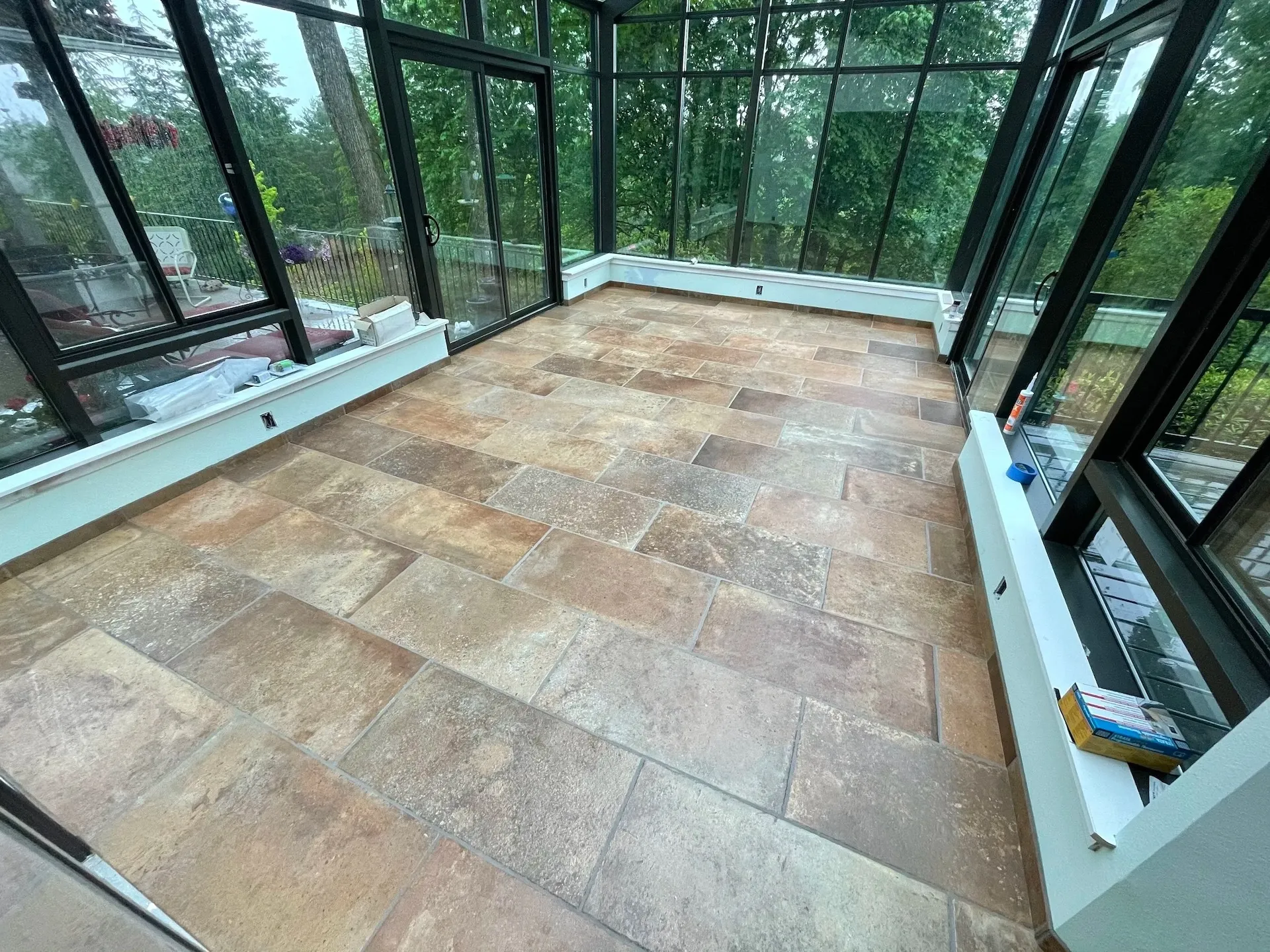 Sunroom or enclosed patio with large black-framed windows and warm stone-look tile flooring overlooking trees