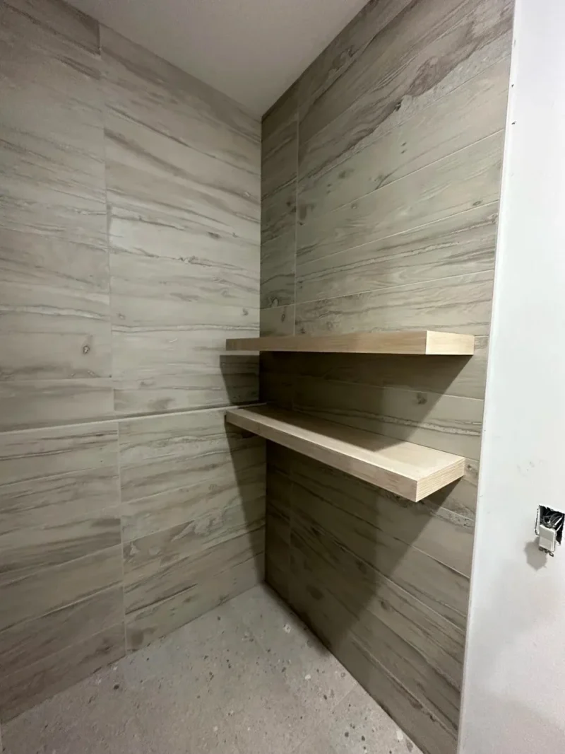 Wood-look tiled shower walls with floating shelves and speckled concrete-style shower floor
