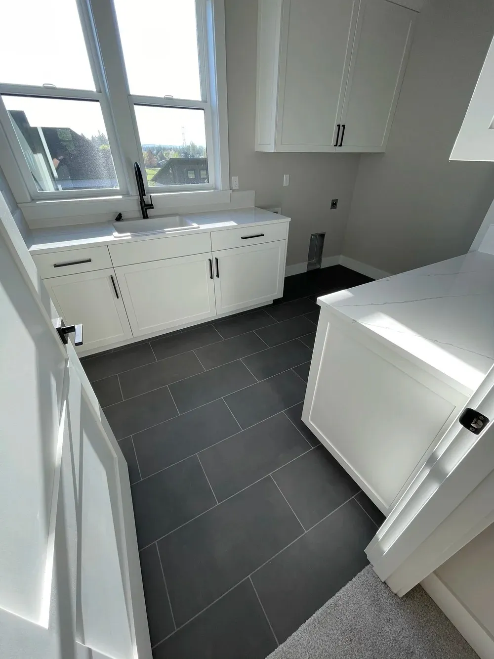 Modern laundry room with white cabinetry, black tile flooring, and large windows