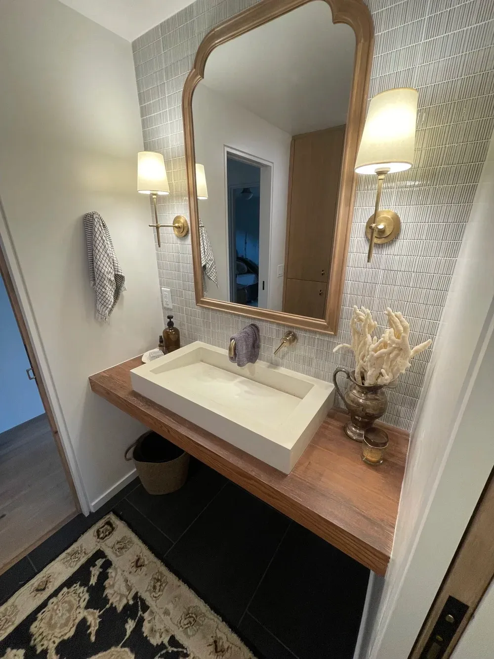 Modern powder bathroom featuring floating wood vanity, textured tile backsplash, and brass sconces