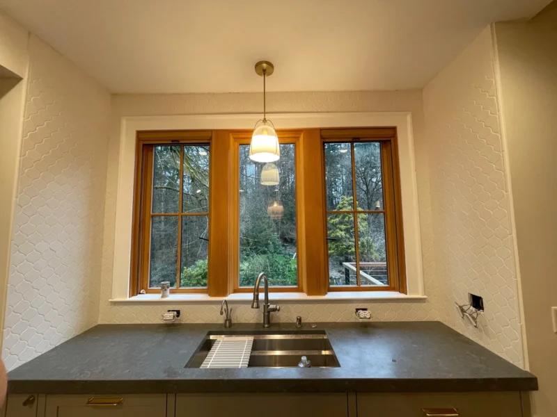 Farmhouse kitchen sink with wood-framed windows, textured backsplash, and dark soapstone countertops