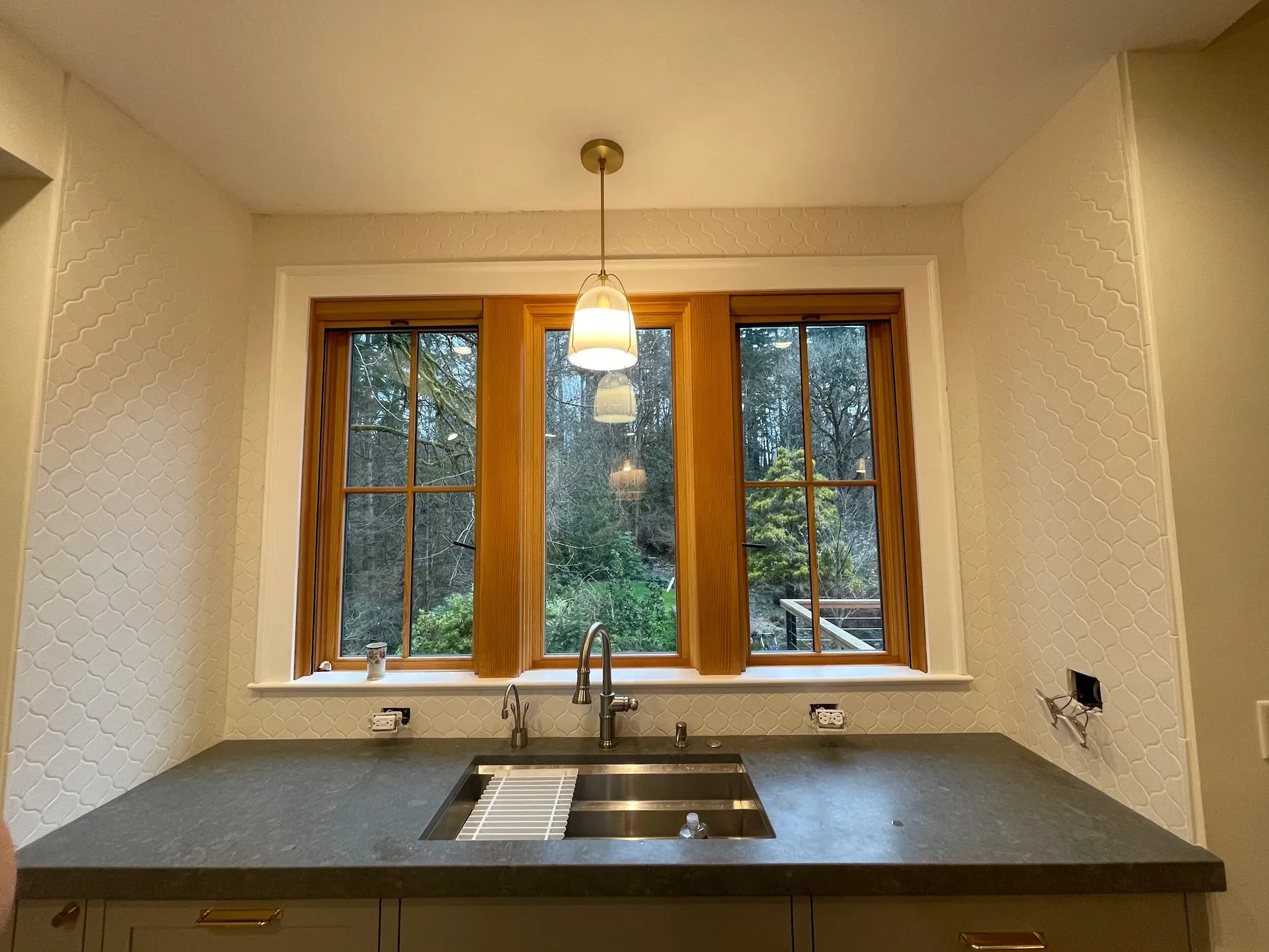 Farmhouse kitchen sink with wood-framed windows, textured backsplash, and dark soapstone countertops